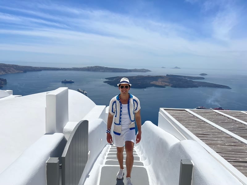 A man wearing a white and blue polo, sunglasses, a white hat, a white tank top and white shorts walking on a stairway in the village of Imerovigli, Santorini, whit the Aegean Sean and the caldera in the background