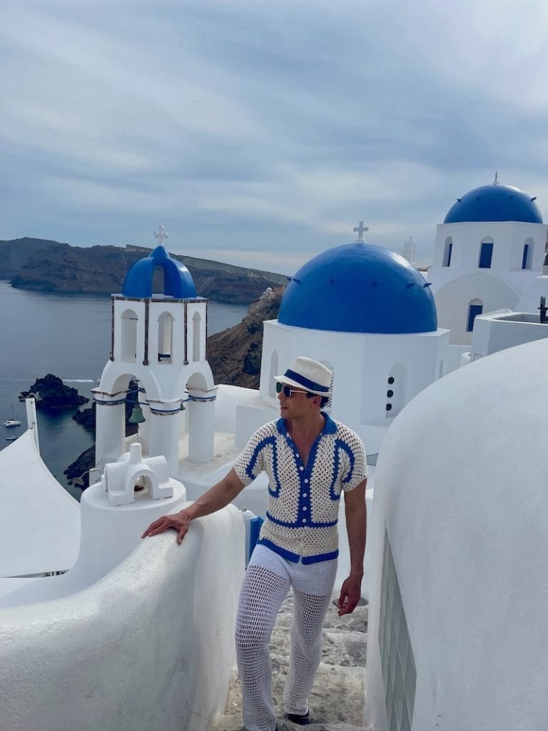 A man wearing a white and blue polo, sunglasses and white trousers walking on a stairway in the village of Oia, Santorini, and three blude-domed churh with white walls in the background